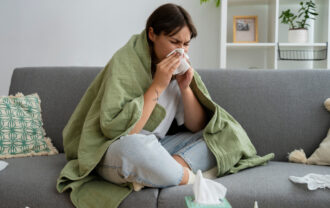 A person wrapped in a green blanket blowing their nose on the couch, surrounded by tissues, suggesting a cold or allergies.