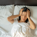 Woman lying in bed with hands on her head, showing signs of discomfort or a headache, possibly struggling with insomnia