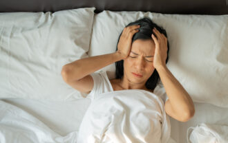Woman lying in bed with hands on her head, showing signs of discomfort or a headache, possibly struggling with insomnia